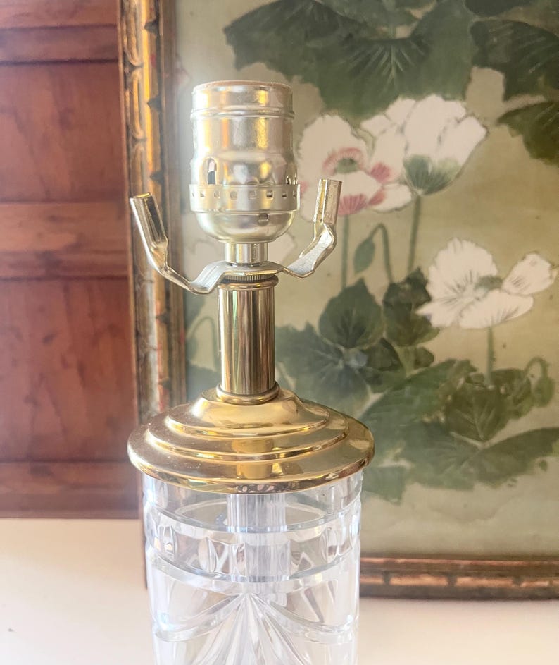 May include: A clear glass lamp base featuring a faceted geometric pattern and a polished brass neck. The top includes a silver-toned metal light socket with two extending arms for shade attachment, showcasing a reflective and transparent design.