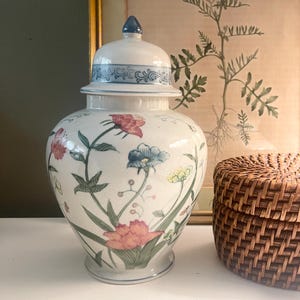 May include: A white ceramic ginger jar with a lid, adorned with a floral pattern in pink, blue, and yellow. The jar has a blue base and a blue finial on the lid. A woven basket is to the right.