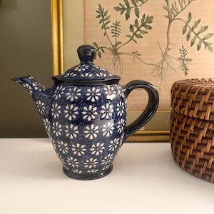 May include: A dark blue ceramic teapot featuring a repeating white floral pattern across its surface. The teapot includes a matching lid with a small knob handle, a curved spout, and a sturdy side handle, all finished with a glossy glaze.