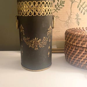May include: A dark gray cylindrical container with gold accents. The top has a decorative gold filigree design. Gold floral and ribbon details adorn the front. Two gold handles are on the sides. A woven basket is to the right.