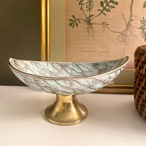 May include: A crescent-shaped bowl with a pale blue and white crackle glaze finish, featuring a gold-toned metallic rim and a matching gold-toned pedestal base. The bowl has a curved, elongated silhouette and sits on a smooth, circular stand.