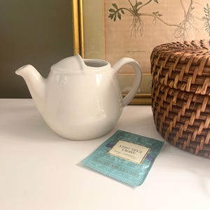 May include: A white ceramic teapot with a curved handle and spout. A teal tea bag with the text "Smoky Earl Grey" is next to the teapot. A woven basket and framed botanical print are in the background.