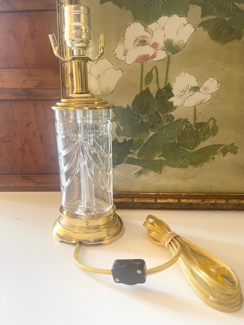 May include: A table lamp featuring a clear, vertically fluted glass body with a decorative fan-like pattern. The lamp has a polished brass base and top fixture, a gold-toned electrical cord, and a black inline switch with a two-prong plug.