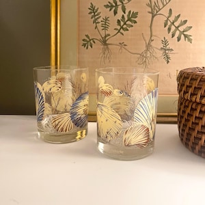 May include: Two clear glass tumblers featuring a decorative wrap-around print of various seashells in shades of blue, gold, and brown, accented with fine sand-like speckles and thin, wispy sea grass illustrations on the exterior surface.