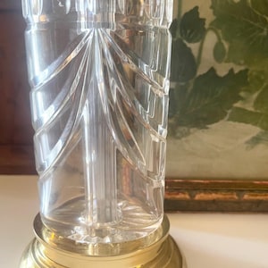 May include: A clear glass lamp base featuring vertical fan-shaped cut patterns and a smooth cylindrical neck. The glass body is mounted on a polished gold-tone circular metal base with a stepped edge, providing a stable foundation for the structure.