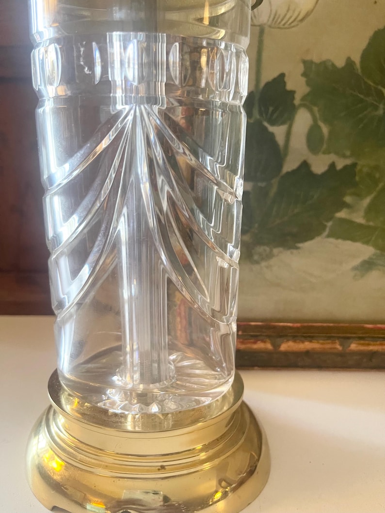 May include: A clear glass lamp base featuring vertical fan-shaped cut patterns and a smooth cylindrical neck. The glass body is mounted on a polished gold-tone circular metal base with a stepped edge, providing a stable foundation for the structure.