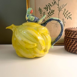 May include: A ceramic teapot with a yellow banana-shaped body and a blue and green handle. A small, colorful parrot figurine sits atop the lid. The teapot is a unique decorative item.