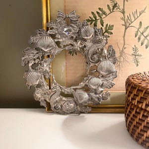 May include: A silver-colored, circular picture frame with a nautical theme. The frame is adorned with detailed seashell, seahorse, and starfish designs. The frame is approximately 10 inches in diameter.