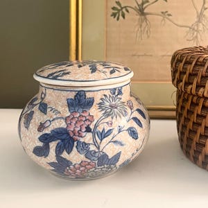 May include: A small, round ceramic jar with a lid, featuring a floral design in pink, blue, and white on a cream background. The jar has a geometric pattern and is likely used for storage or decoration.