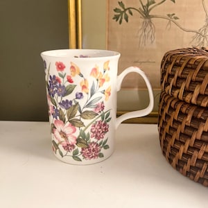 May include: A white ceramic mug featuring a colorful botanical print of various wildflowers including pink, purple, and yellow blossoms with green leaves. The mug has a smooth, cylindrical body with a curved handle on the right side and a flared rim.
