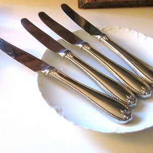 May include: Five silver-colored butter knives arranged on a white, scalloped-edge plate. The knives have a decorative rope-like design on the handles and a simple, rounded blade. The knives are angled towards the upper right.