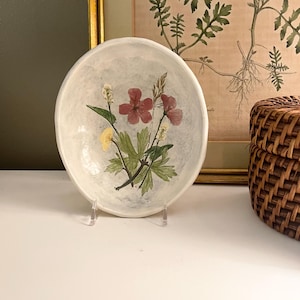 May include: A round, off-white ceramic bowl featuring a hand-painted botanical illustration in the center. The design depicts a cluster of small flowers with pink, yellow, and white petals, accompanied by green leaves and stems against a light gray wash.