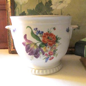 May include: A white ceramic planter with a floral design featuring purple, orange, and blue flowers. The planter has two small handles and a gold trim. The base has a decorative, ridged design.