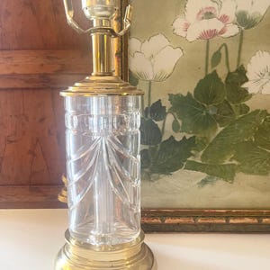 May include: A table lamp base featuring a clear glass cylindrical body with vertical etched lines and a scalloped pattern. The lamp has a polished brass circular base, a matching brass neck, and a metal socket assembly at the top for a light bulb.