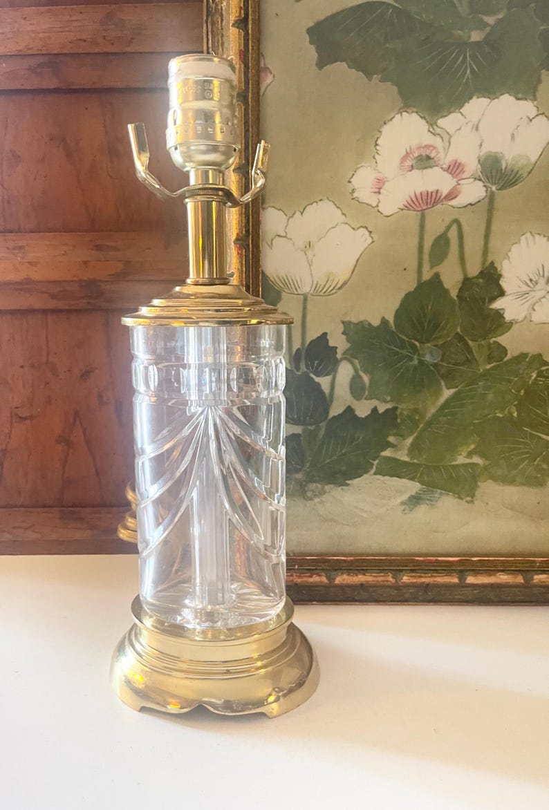 May include: A table lamp base featuring a clear glass cylindrical body with vertical etched lines and a scalloped pattern. The lamp has a polished brass circular base, a matching brass neck, and a metal socket assembly at the top for a light bulb.