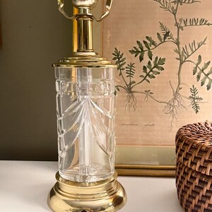 May include: A table lamp base featuring a clear, cylindrical glass body with vertical cut patterns. The lamp has a polished brass circular base and a matching brass top fitting with a standard bulb socket and two metal shade support arms.