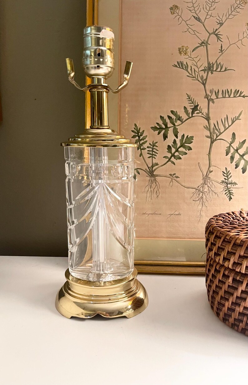 May include: A table lamp base featuring a clear, cylindrical glass body with vertical cut patterns. The lamp has a polished brass circular base and a matching brass top fitting with a standard bulb socket and two metal shade support arms.