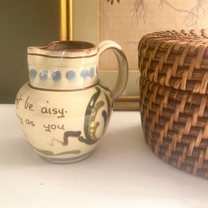 May include: A small, off-white ceramic pitcher with a handle and spout. The pitcher is decorated with blue, green, and brown painted designs and the words "t be aisy. y as you". A woven basket is in the background.