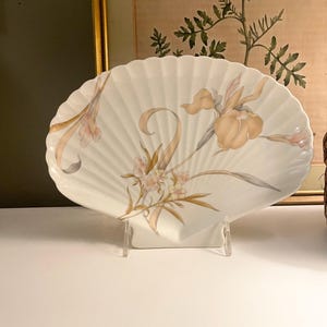 May include: A white ceramic serving dish shaped like a large scallop shell, featuring a delicate painted design of soft orange and yellow iris flowers with green stems and leaves, displayed upright on a clear plastic stand for decorative viewing.