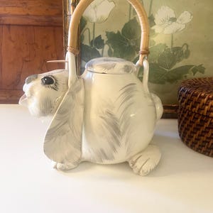 May include: A white ceramic teapot shaped like a rabbit, with gray brushstroke details and a bamboo handle. The teapot has a lid and is sitting on a white surface. The teapot is a decorative item.