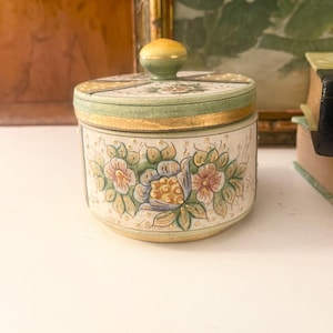 May include: A round, hand-painted ceramic jar with a lid. The jar is decorated with floral designs in shades of pink, blue, and yellow on a cream background. The lid has a green and gold border and a small yellow knob. The jar is likely used for storage.