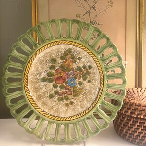 May include: A round ceramic plate with a sage green openwork lattice rim and a central cream-colored field featuring a hand-painted floral motif in shades of orange, blue, and green, bordered by a yellow and brown rope-patterned decorative band.