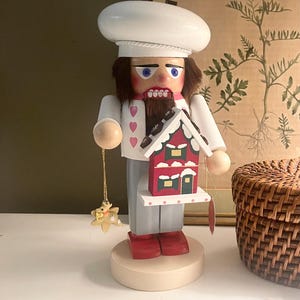May include: A wooden nutcracker in the shape of a chef, wearing a white hat and uniform, holding a red and green house ornament. The nutcracker has a brown beard and is standing on a beige base. A gold star-shaped ornament hangs from his hand.