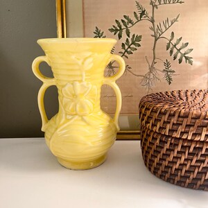 May include: A bright yellow ceramic vase featuring a glossy finish, a flared rim, and two curved handles on the sides. The body of the vase is decorated with a raised, molded floral pattern and horizontal ribbed detailing around the neck and base.