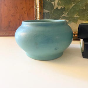 May include: A round, light blue ceramic vase with a wide body and a small base. The vase has a smooth, matte finish and a slightly uneven color distribution, creating a subtle visual texture. The vase is a decorative item.