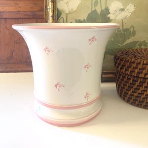May include: White ceramic vase with a flared top and a pink rim and base. The vase features small pink floral designs scattered across the surface. A decorative piece for the home.