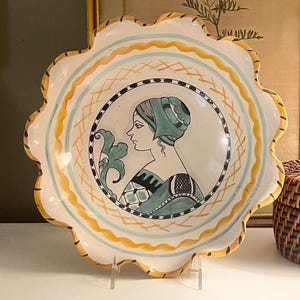 May include: A scalloped ceramic plate featuring a central circular illustration of a person in profile wearing a green head covering and patterned clothing. The plate has a white background with yellow and light blue decorative border patterns.