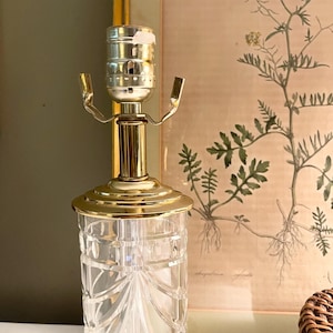 May include: A table lamp base featuring a clear, cylindrical glass body with vertical cut-glass detailing and a gold-toned metal neck and socket assembly. The lamp has a polished gold-colored circular base plate and a standard metal light bulb socket.