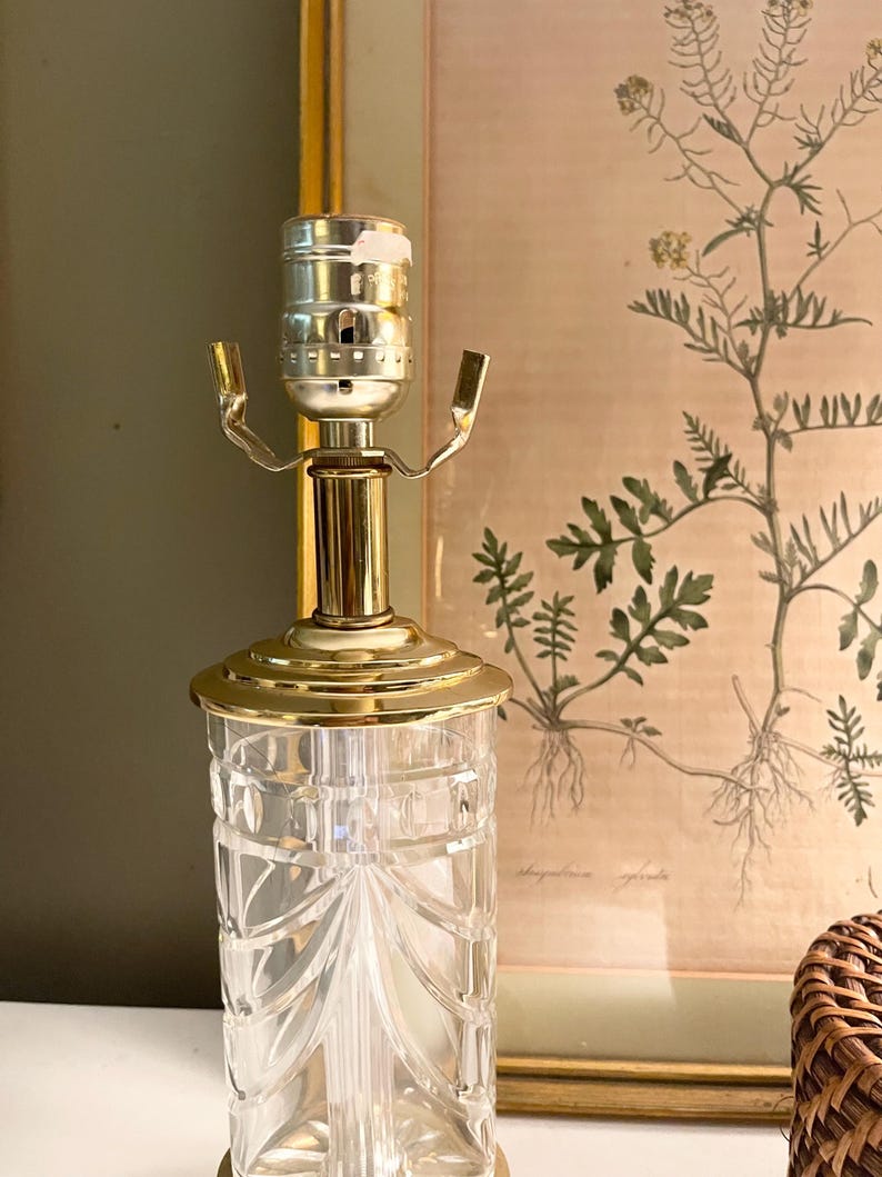 May include: A table lamp base featuring a clear, cylindrical glass body with vertical cut-glass detailing and a gold-toned metal neck and socket assembly. The lamp has a polished gold-colored circular base plate and a standard metal light bulb socket.