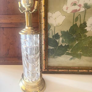 May include: A table lamp featuring a clear, cylindrical glass body with vertical cut patterns, set upon a polished brass base. The lamp includes a brass neck and a metal socket assembly at the top, designed to hold a light bulb without a shade.