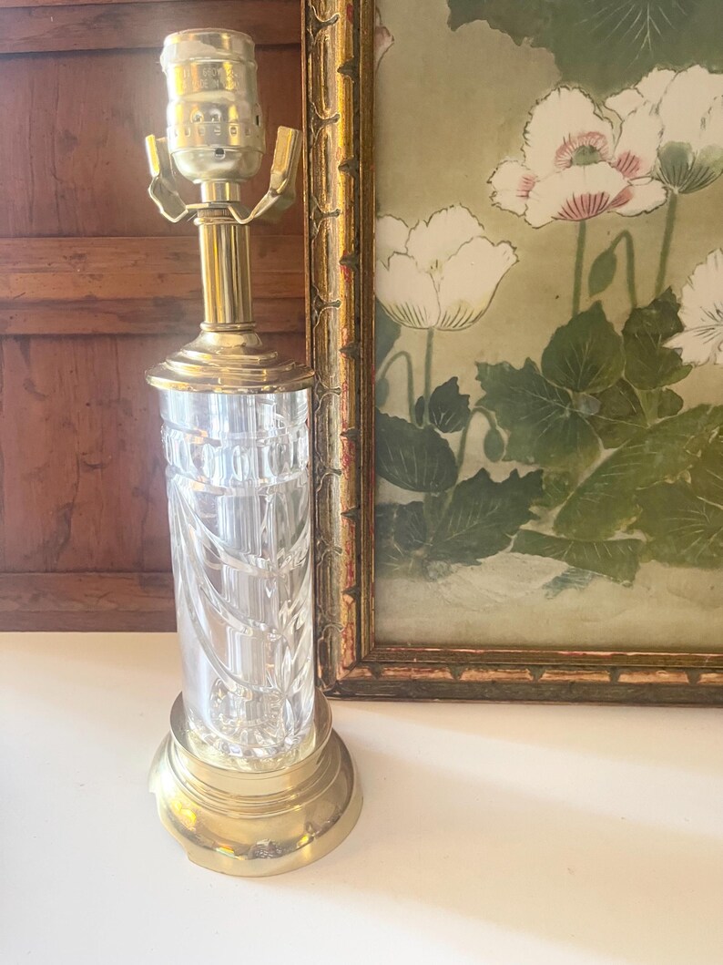 May include: A table lamp featuring a clear, cylindrical glass body with vertical cut patterns, set upon a polished brass base. The lamp includes a brass neck and a metal socket assembly at the top, designed to hold a light bulb without a shade.