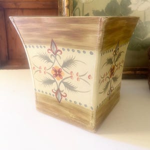 May include: A square, decorative wooden planter with a cream-colored background and floral design. The planter has a brown border and is painted with a symmetrical floral pattern in shades of red, orange, and gray.