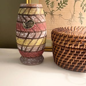 May include: A vintage, hand-decorated ceramic vase with a cylindrical shape. The vase features horizontal bands of red, yellow, and white, with a diamond pattern. The vase has a small base and a label that reads "Rossini Originals Hand Decorated Italy."