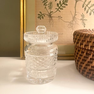 May include: A clear, cut-glass jar with a lid, featuring a diamond pattern on the body and a decorative knob on top. The jar is sitting on a white surface. A woven basket and a framed botanical print are in the background.