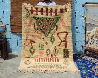 May include: Cream-colored, hand-woven rug with geometric designs in red, green, and pink. The rug features a central triangle and various diamond and rectangular shapes. The rug has a fringed edge and is held up by a person.