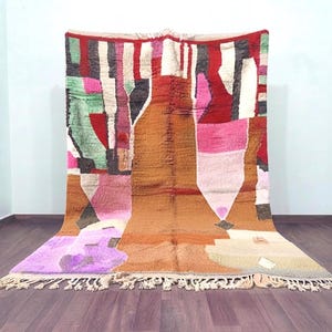 May include: A large rectangular area rug featuring a vibrant abstract pattern with blocks of brown, red, pink, green, black, and cream. The rug has a thick pile texture and is finished with a fringe of knotted yarn along the bottom edge.