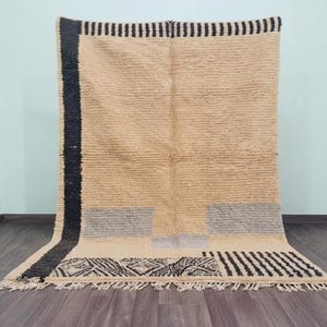 May include: A rectangular area rug featuring a beige base with a thick black vertical stripe on the left side. The design includes gray rectangular blocks, black geometric patterns, and black horizontal stripes at the top and bottom edges with fringe.