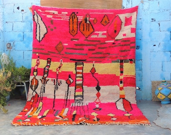 May include: A vibrant pink rug with geometric patterns in orange, yellow, black, and white. The rug features a central design with vertical and horizontal stripes, and abstract shapes. The rug is displayed against a blue brick wall.