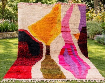 May include: A rectangular area rug featuring a cream-colored background with abstract shapes in vibrant shades of yellow, pink, purple, red, black, and olive green. The rug has a thick, textured pile and includes knotted fringe along the top and bottom edges.