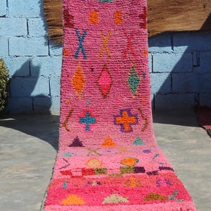 May include: A bright pink rug featuring colourful geometric designs in orange, teal, purple, and yellow. The rug has a textured, woven appearance with a fringe along the bottom edge. The design includes diamond, cross, and triangle shapes.