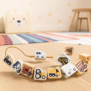 Könnte beinhalten: Ein Holzperlenspielzeug mit verschiedenen Fahrzeugdesigns, darunter ein Auto, ein LKW und ein Bus. Die Perlen sind in sanften Farben wie Weiß, Gelb und Braun bemalt und mit einer naturfarbenen Schnur verbunden. Das Spielzeug ist für Kinder.