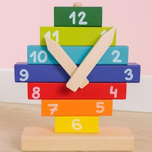 May include: A colorful wooden clock toy with a rainbow of blocks numbered 1-12. The clock hands are made of light wood. The clock sits on a natural wood base. The background is a light pink.