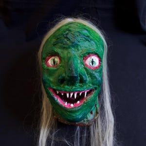 May include: A grotesque, green-skinned mask with white hair and a wide, toothy grin. The eyes are red with white highlights. The mask is likely for Halloween or a similar event.