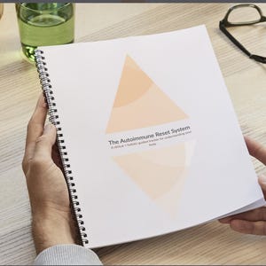 May include: A spiral-bound notebook with the title "The Autoimmune Reset System" and a geometric design in shades of orange. The notebook is held open, revealing the pages. A glass of water and eyeglasses are visible in the background.