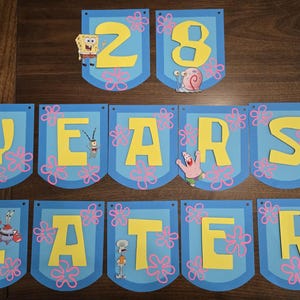 Op de afbeelding: Een set blauwe banners met gele letters en roze bloemaccenten. De banners lezen "28 YEARS LATER" met cartoonfiguren uit de SpongeBob SquarePants-serie. Een feestelijke decoratie.