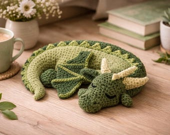 May include: A handmade crochet dragon in shades of green, with cream-colored horns and closed eyes, resting on a wooden surface. The dragon has detailed wings and a textured body. The scene includes a cup of coffee, flowers, and books.